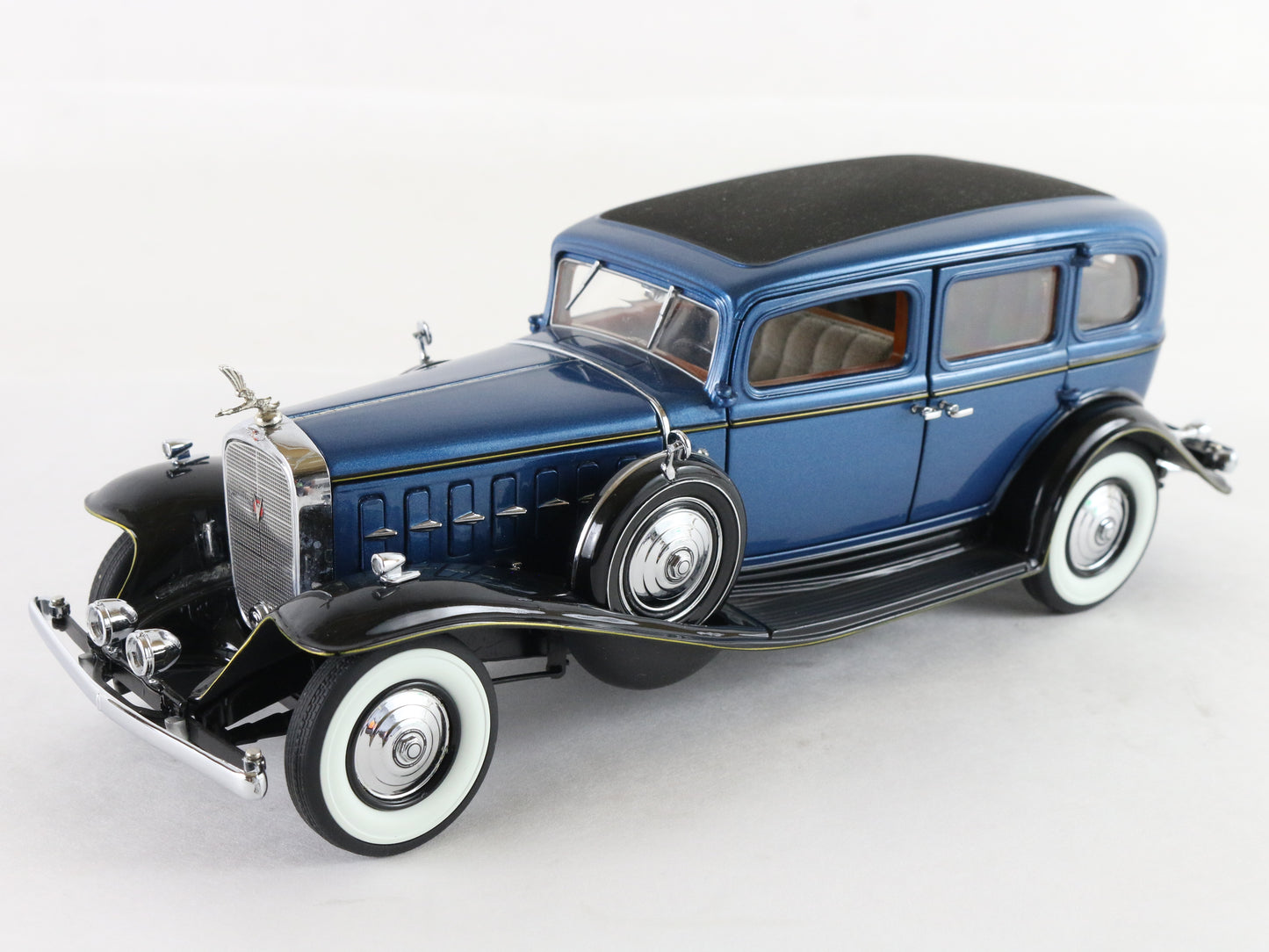 A vintage-style blue model car with black fenders, black roof, white-walled tires, and chrome details, photographed on a plain light background.